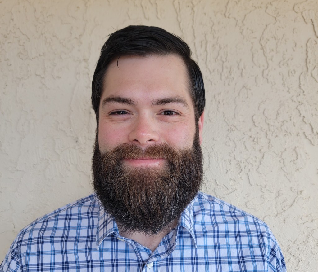Close-up of a smiling man with a full beard, wearing a blue and white checkered shirt, against a textured beige background.